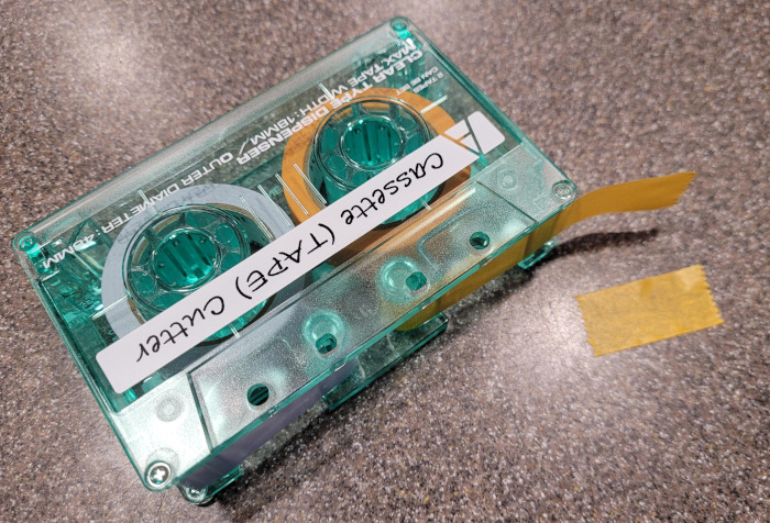 A teal translucent plastic washi tape roll dispenser that looks like an audio cassette. A piece of orange tape is being dispensed.