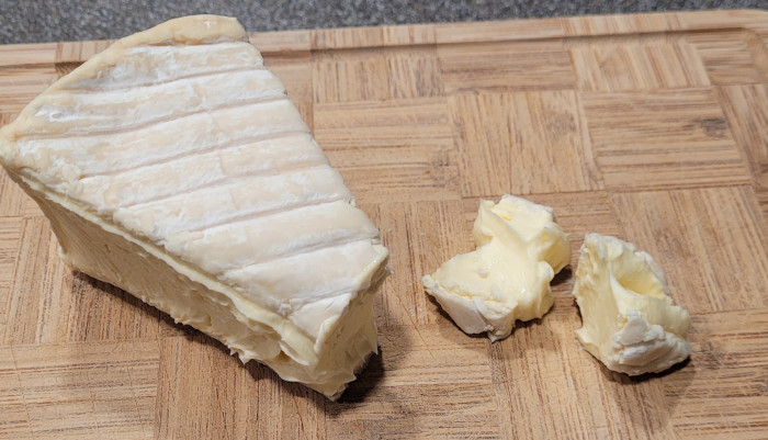 Photo of cheese on cutting board. It has a white rind with stripes. Two chunks have been severed from the wedge.